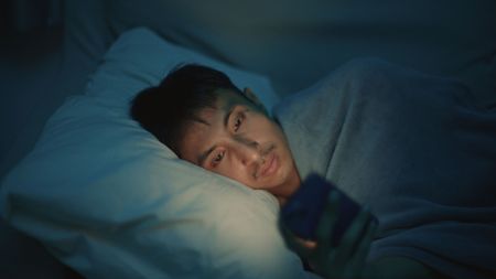 Asian man using smartphone on bed at night, insomnia and social media addiction.