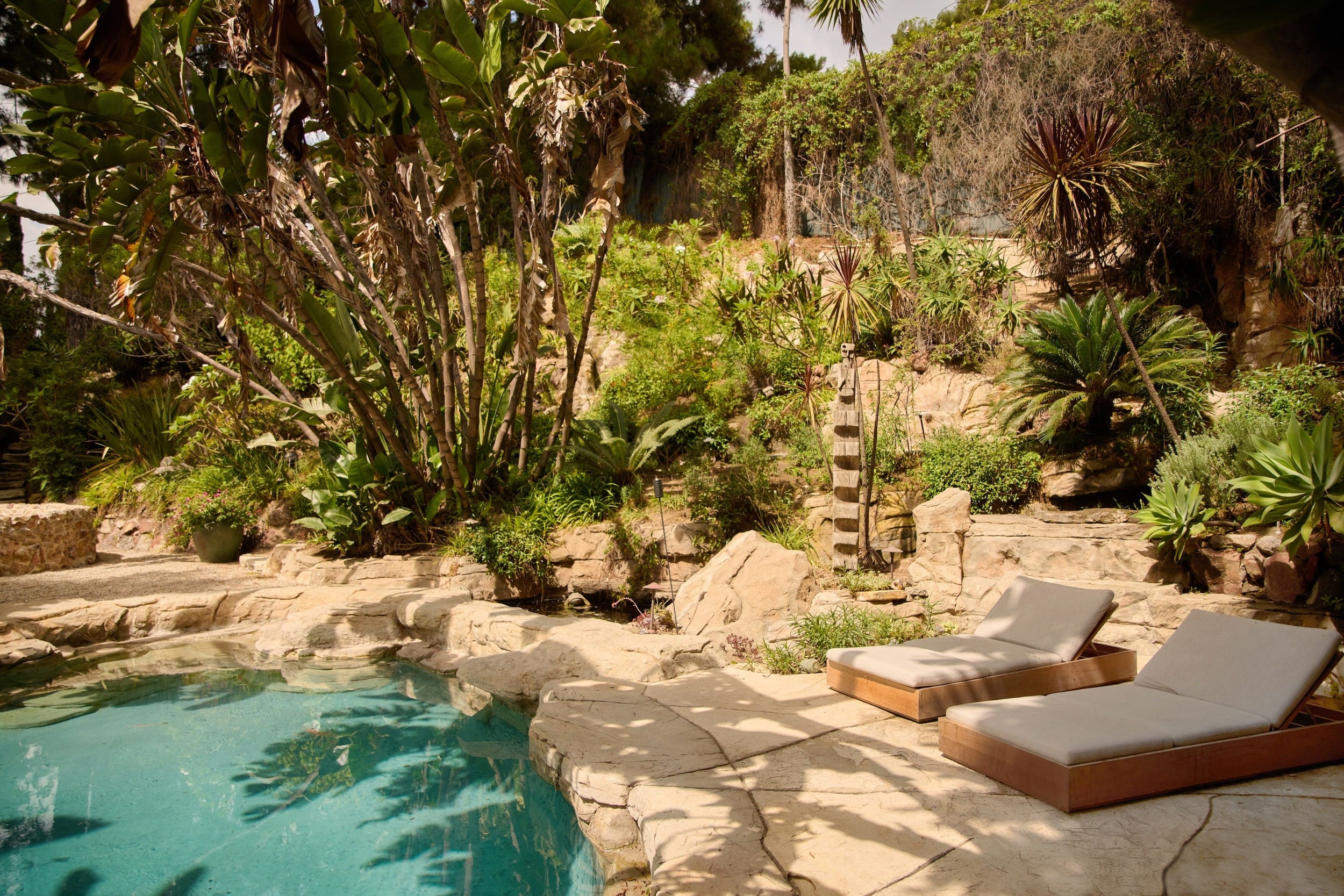corner of pool surrounded by rocky surface and layers of greenery, two white deckchairs facing the water