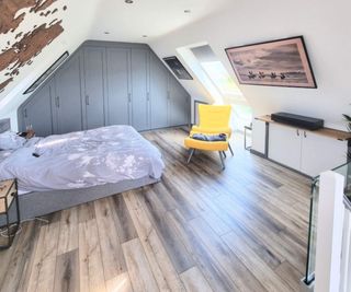 A new bedroom within a loft conversion, with built-in grey wardrobes to one end and a staircase to the other. The room also features a TV on the sloping ceiling, a double bed and a yellow chair.