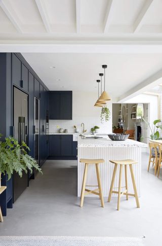 Image of a white, open-space kitchen with tall navy blue cabinets. There is a white kitchen island in the middle of the room with three pendant lights hanging over it. There are live plants around the space.