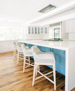 A white kitchen with white cabinets on the walls, a white kitchen island with light blue cabinets underneath, and four white rattan stools next to it, with rich brown wooden flooring underneath