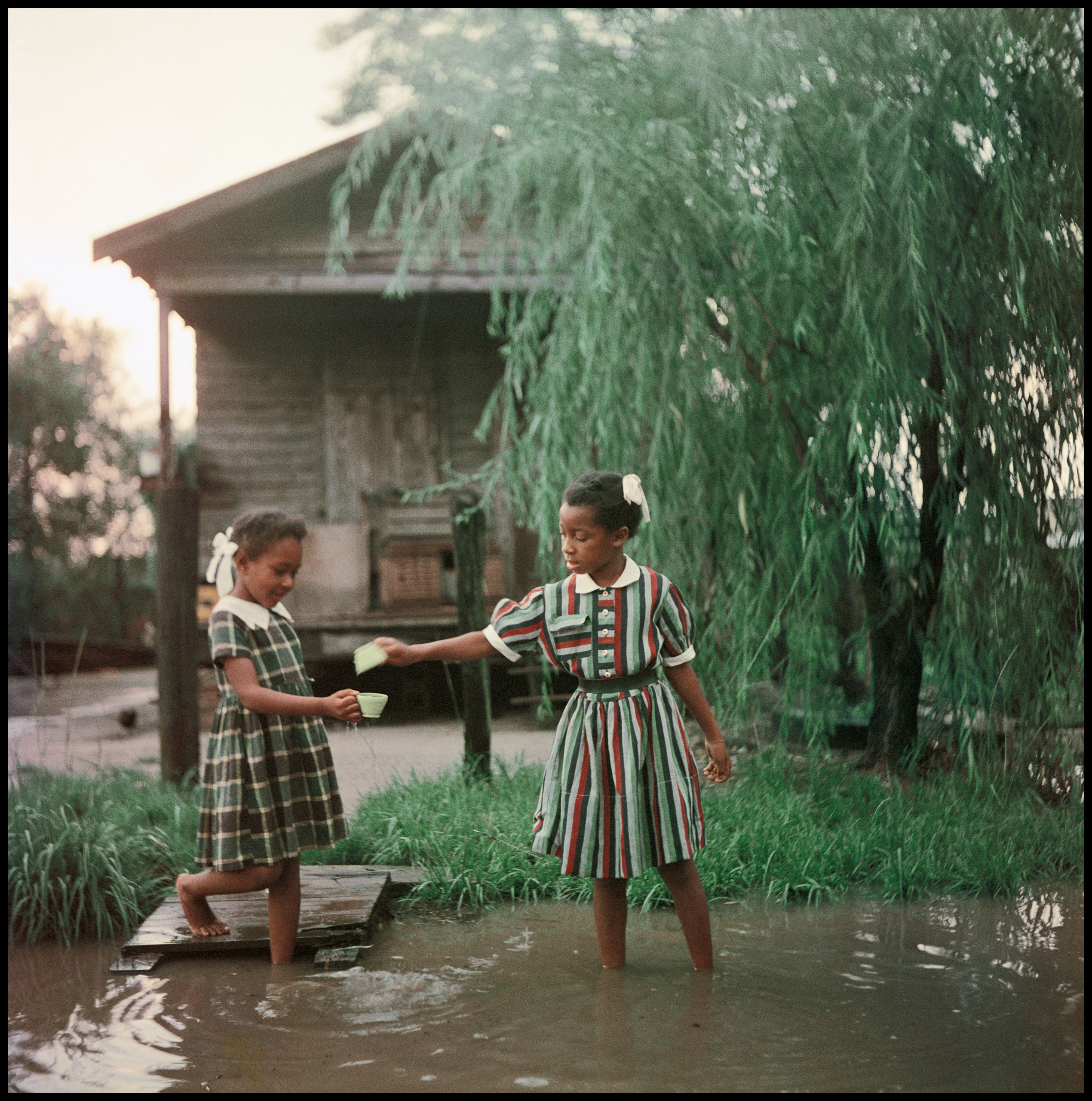 Two young girls in matching striped and checked dresses stand barefoot in floodwater beside a wooden house, passing a small cup between them