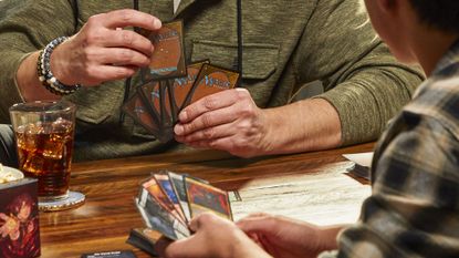 Two people hold Magic: The Gathering cards, seated opposite each other on a table