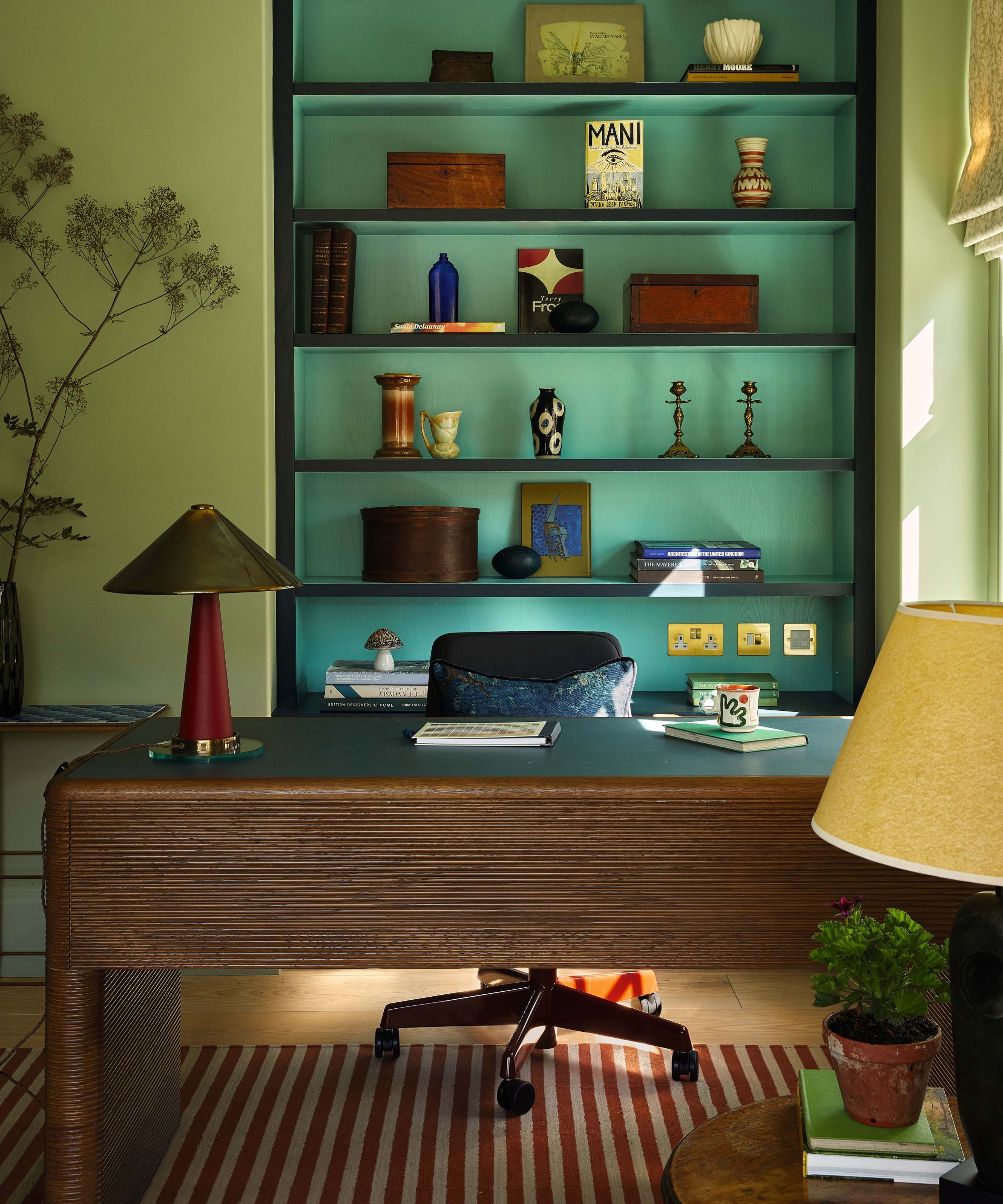 a pale green colorful home office with a ribbed dark wooden desk, bookcase full of trinkets and a red striped rug