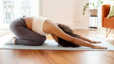 woman resting in child's pose on a mat in a living room setting, sideways to the camera.