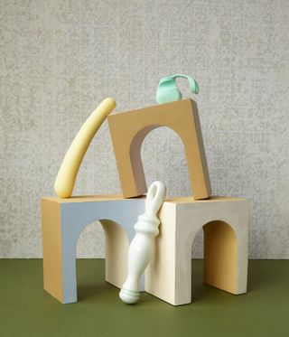 Grey textured background, olive green base, three small wooden arch blocks, three sex toys displayed on and around the blocks