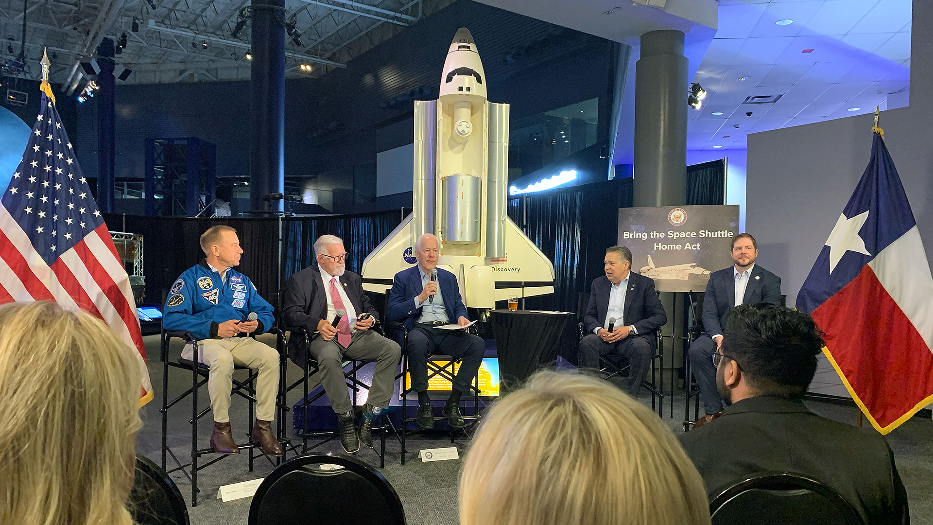 four men in business suits and one man in a NASA astronaut blue flight jacket sit in front of a large model of a space shuttle with U.S. and Texas flags flanking them.