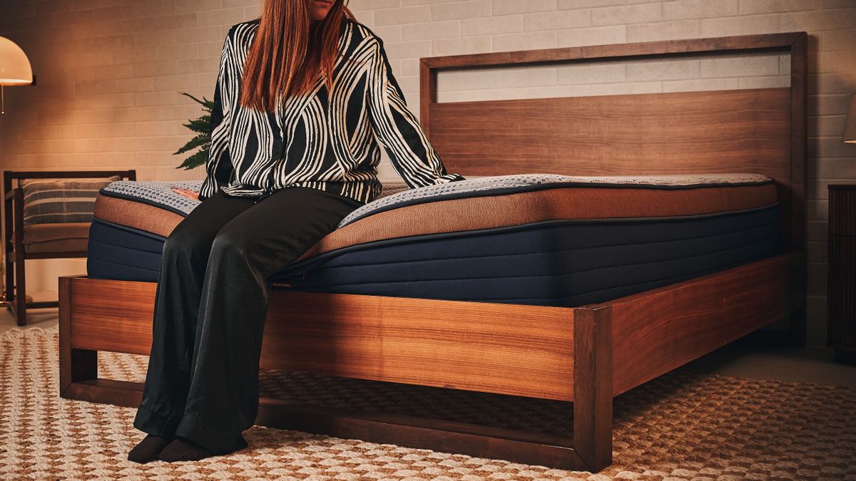A woman in black and white silk pyjamas sitting on edge of Brooklyn Bedding Memory Foam Mattress on wooden bedframe in warmly lit sleep studio