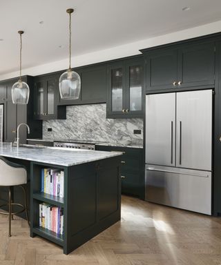 A kitchen with black cabinets, a kitchen island with a gray marble countertop and books underneath it and two glass domed pendant lights above it