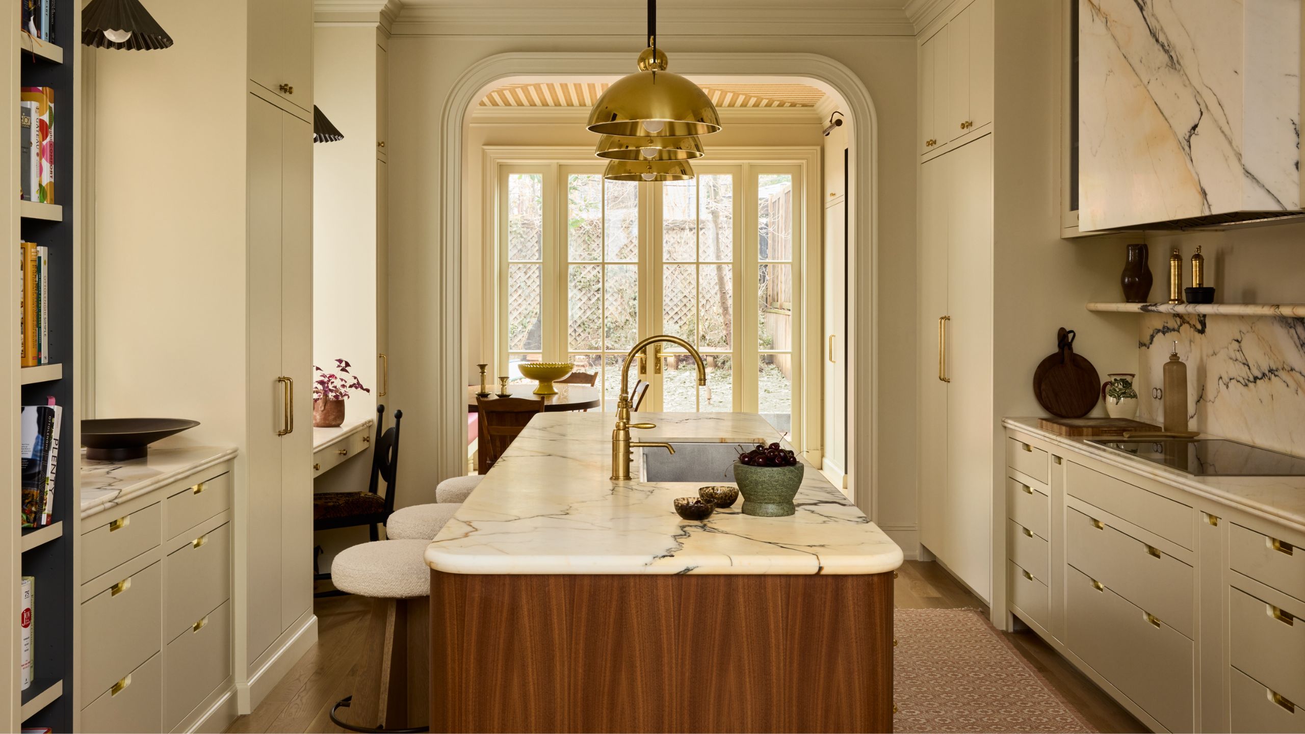 a layered warm neutral kitchen with statement marble counters and cooker hood, with a dark wooden curved island styled with brass pendants and an arched doorway looks out to the back
