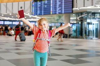 young child standing in departure lounge with arms up and holding her passport