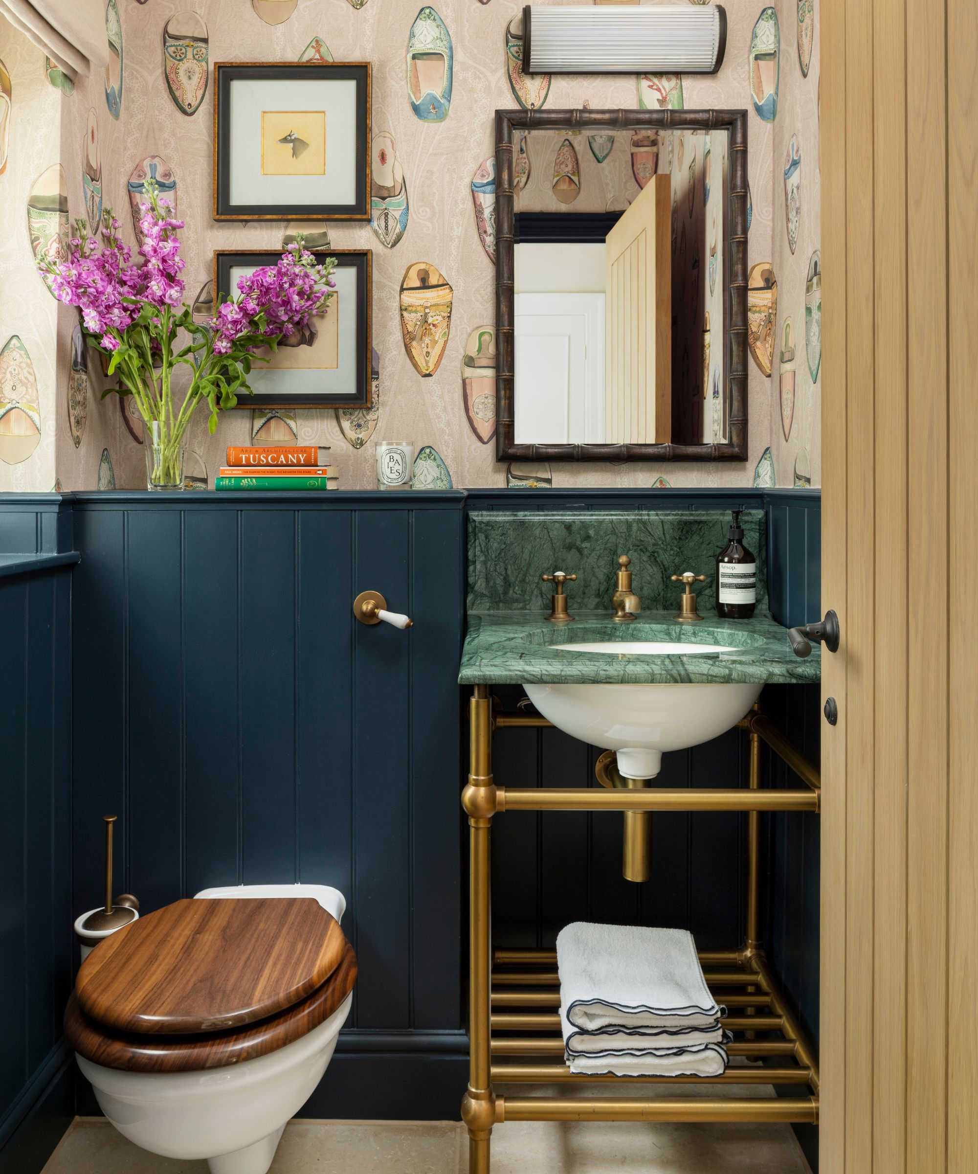 a small powder room with navy blue half height paneling, wallpaper, a small brass vanity with green marble sink and vintage art and mirror