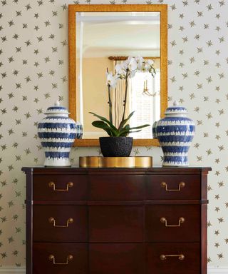 A console table with two symmetrical blue and white vases, a plant and a gold mirror, all against a starry wallpaper