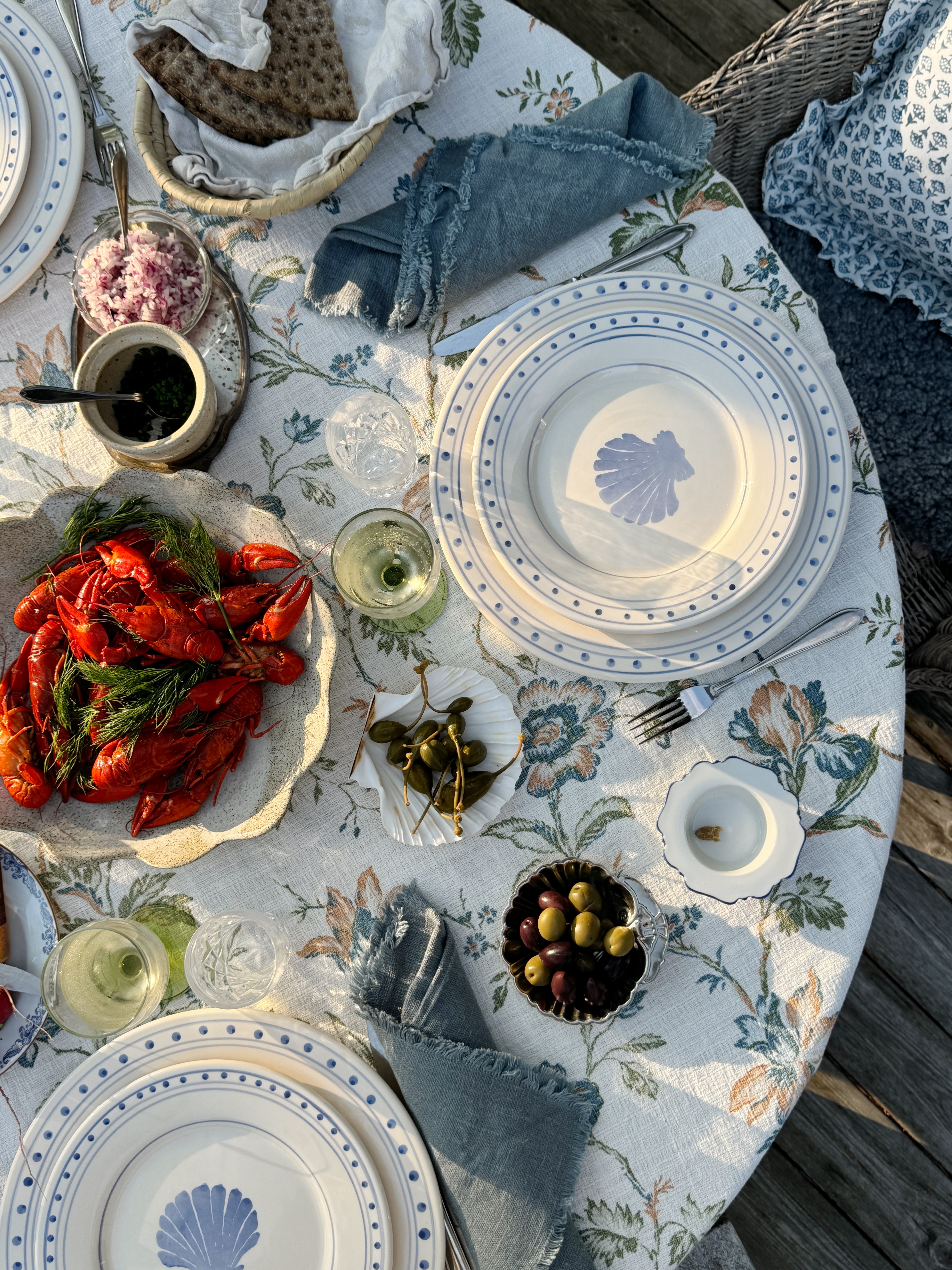 A coastal themed, outdoor dining table setting with a floral patterned, white table cloth and shell printed plates.