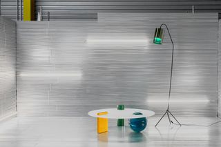 The round deep green, rectangular orange, and ball-shaped blue volumes are supporting the white circular tabletop and emerge on top of it. A floor lamp in black, with a green light, stands next to it.
