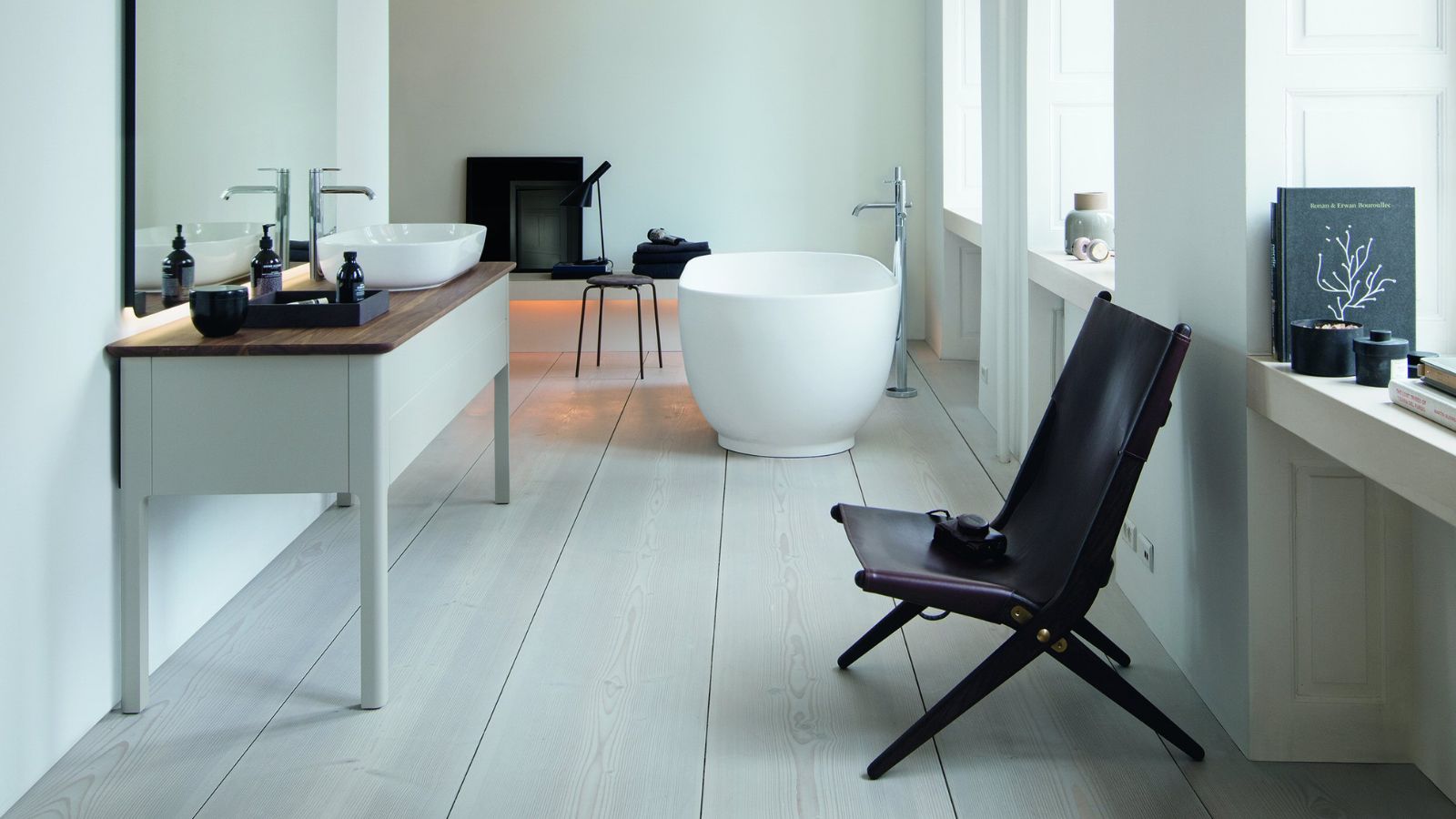 A spacious, minimal Scandinavian bathroom with white walls, wide pale ash floorboards and tall windows. A white console vanity unit with a walnut worktop and twin vessel basins sits beneath a lit mirror, while a sculptural white freestanding bath with a chrome floor-mounted tap occupies the far end.