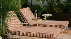 Image of two, striped outdoor lounge chairs with a white metal frame. There are matching white side tables between the chairs, and there are green plants all around.