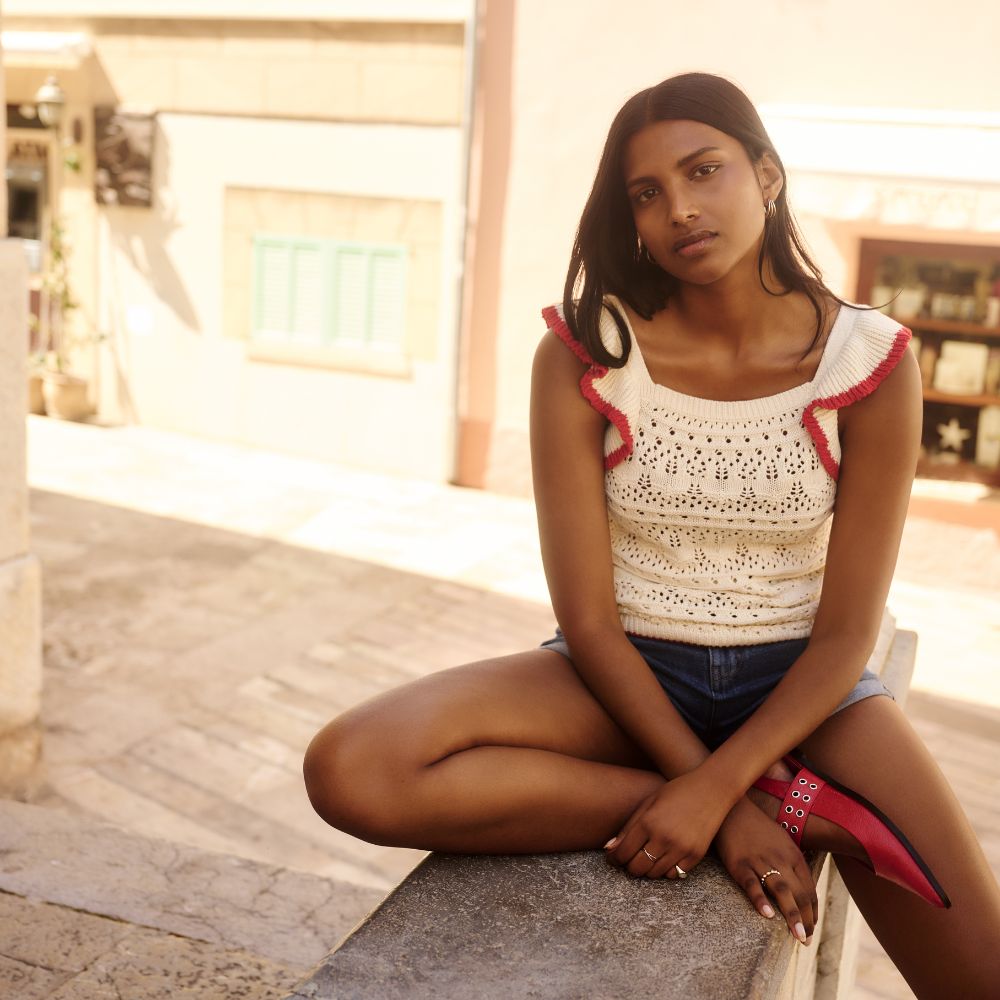 Model sat on a wall wering a white and red crochet top, jean shorts and red ballet flats sold at Simply Be
