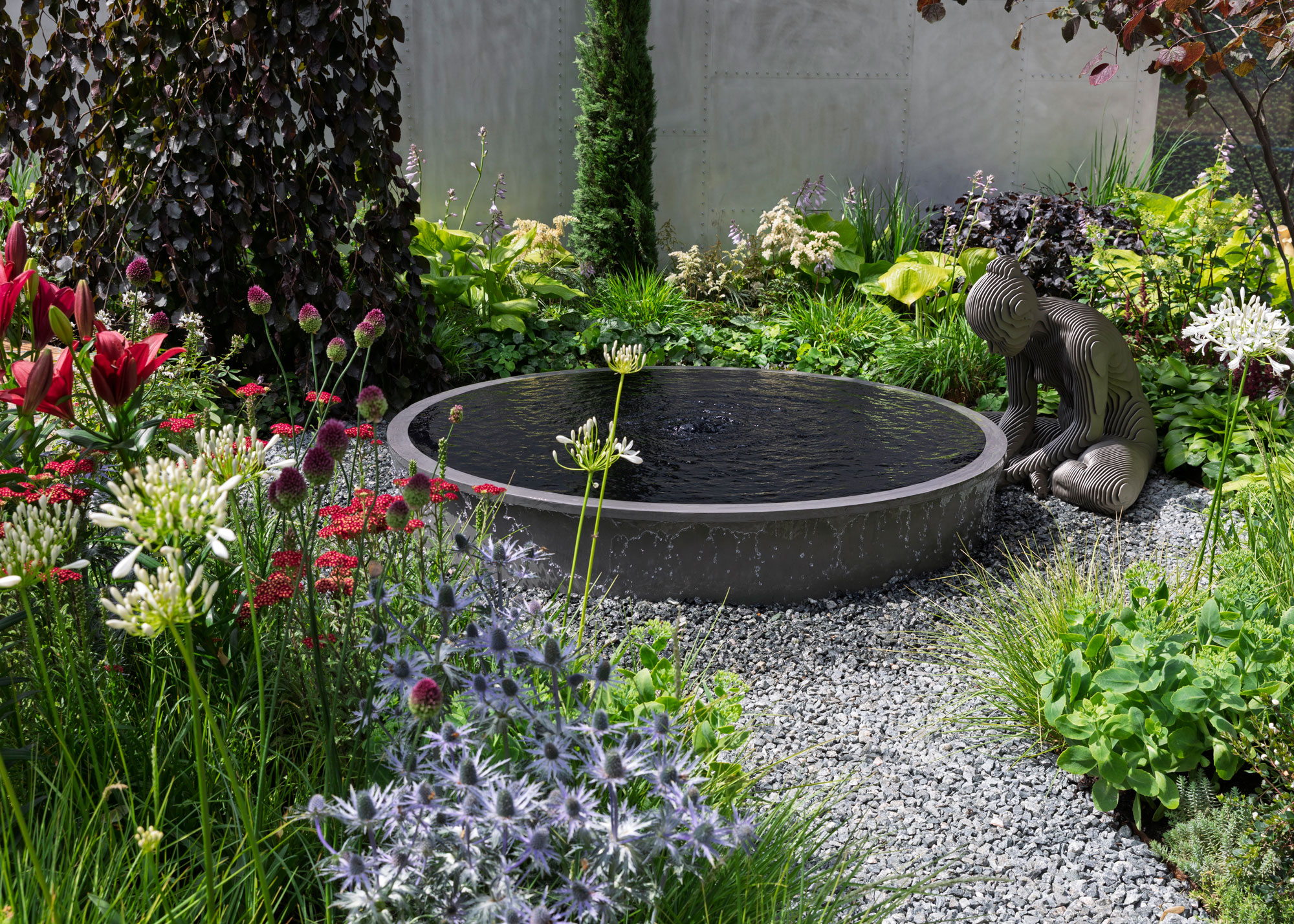 gravel garden with water feature and garden sculpture of woman, with colourful planting including red lilies