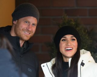 Prince Harry and Meghan Markle wearing winter coats and hats smiling and looking to the right