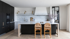 a large classic but modern kitchen with black cabinets and kitchen island and white marble counters and splash back. 2 woven bar stools tuck under the island and a large vase of flowers sits on the counter