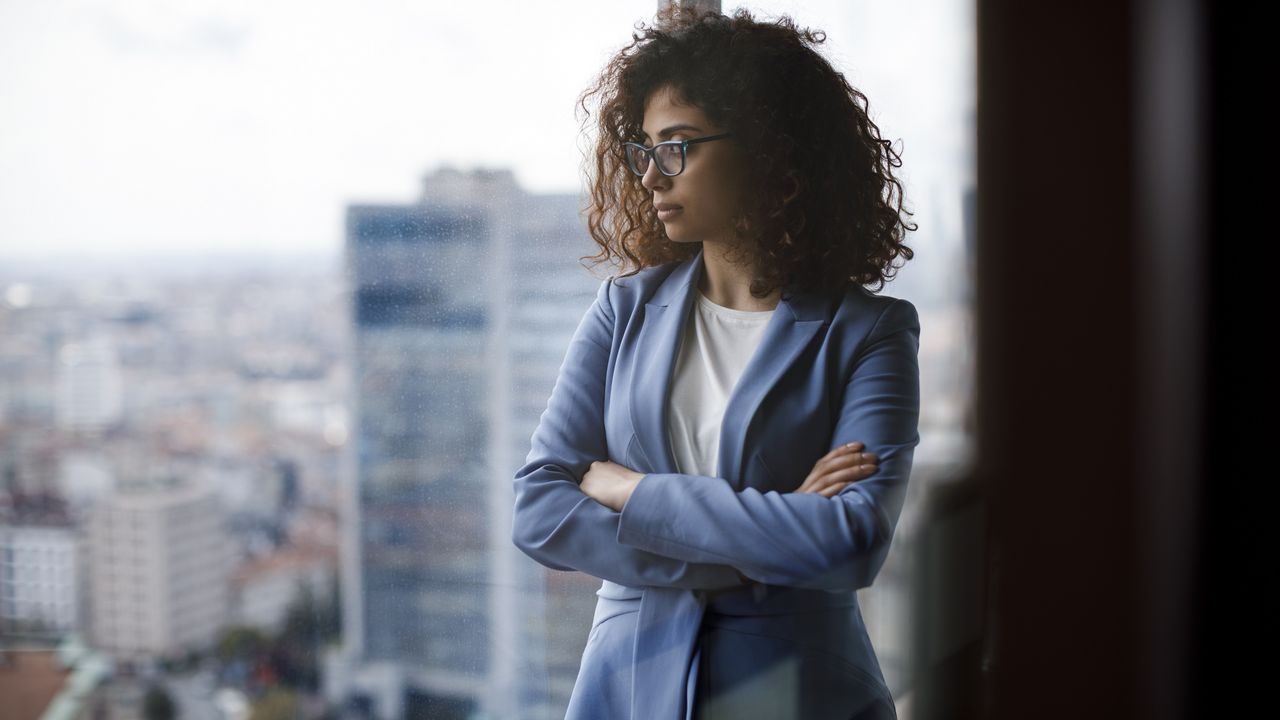 A woman stands with her arms crossed next to a window in a high-rise office building.