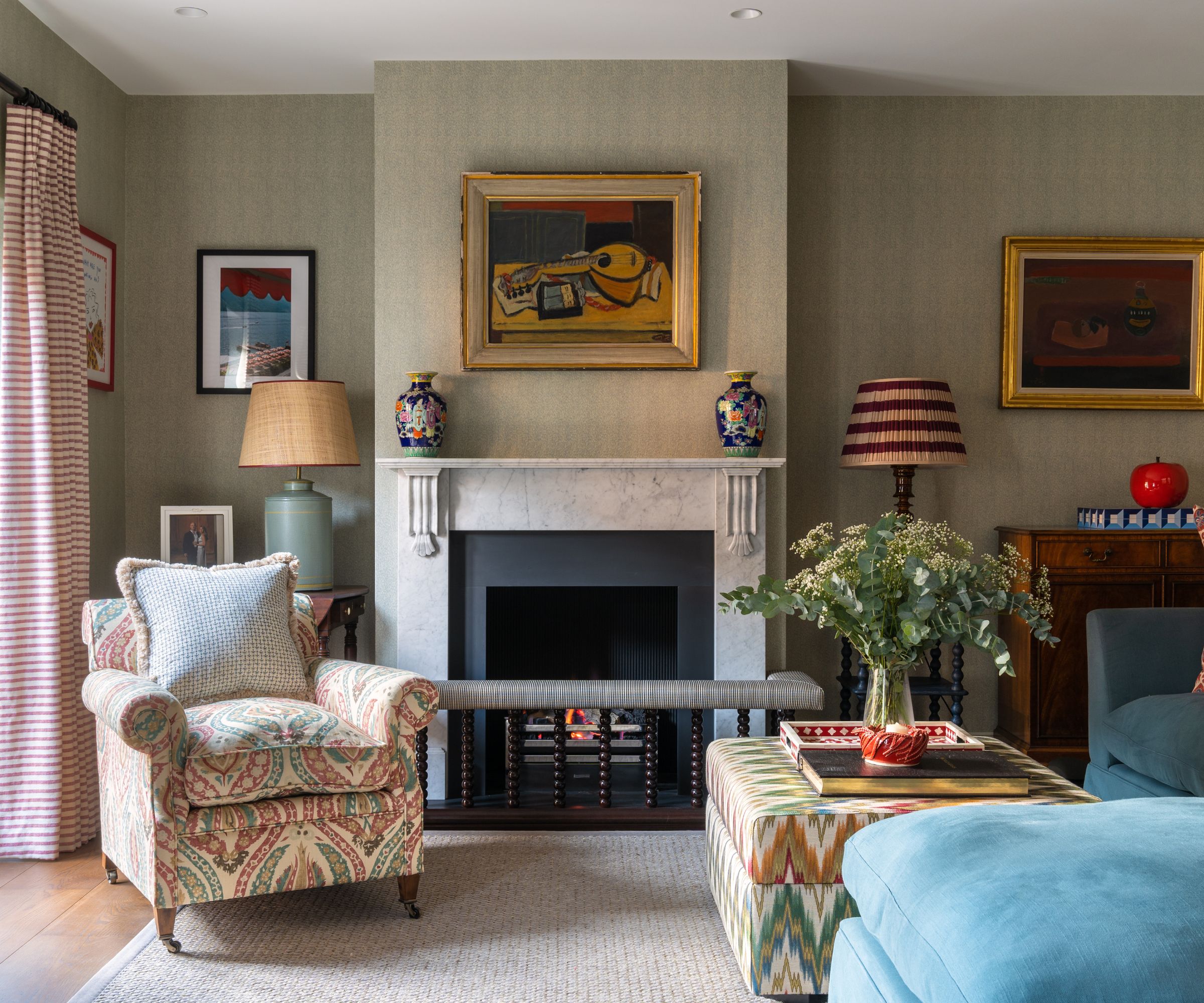 Living room with impressionist art above fireplace, upholstered armchair, fireplace and blue velvet sofa and ottoman with plant