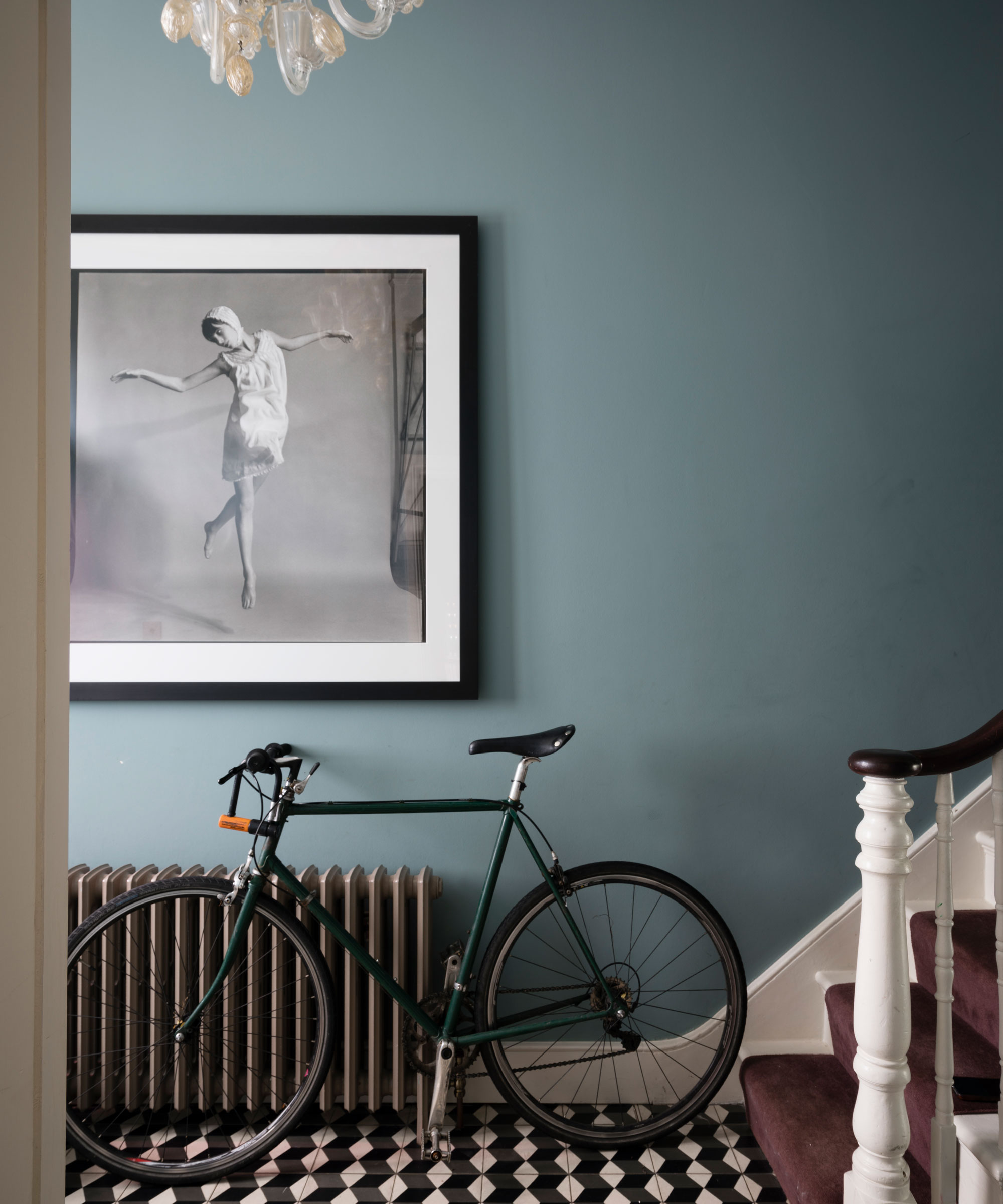 an elegant hallways in a modern home, with black and white tiled floor, a blue wall, a traditional staircase with white and brown bannister, a bicycle up against the wrought-iron radiator, and a painting of a dancing girl hanging on the wall