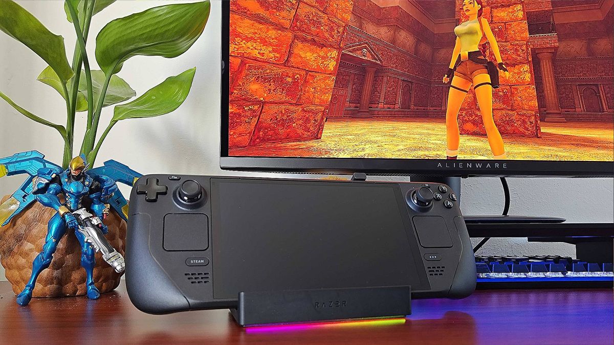 Razer Handheld Dock with Steam Deck sitting on cradle, pink and yellow RGB lighting on, and Alienware monitor in background with Tomb Raider Trilogy gameplay on screen. 