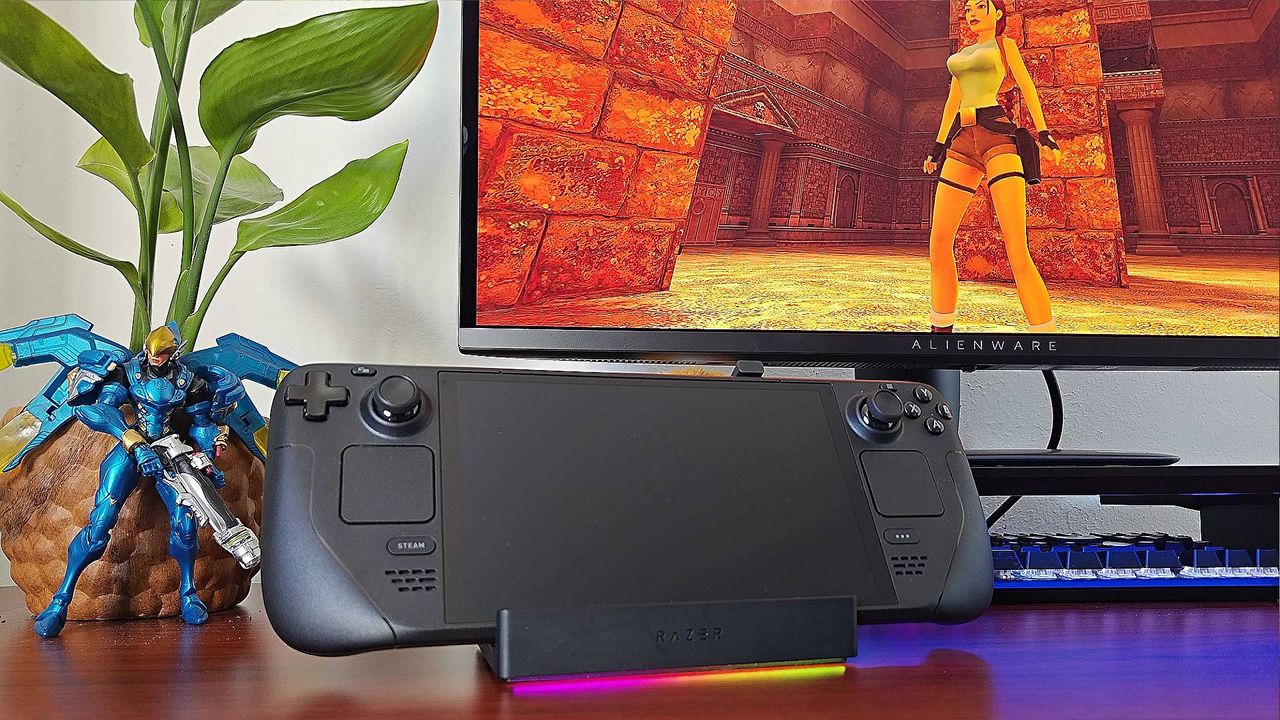Razer Handheld Dock with Steam Deck sitting on cradle, pink and yellow RGB lighting on, and Alienware monitor in background with Tomb Raider Trilogy gameplay on screen.