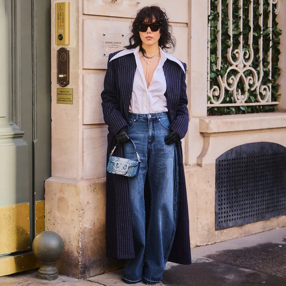 woman wears white button down, pinstripe duster coat, wide-leg jeans, and a mini denim valentino bag