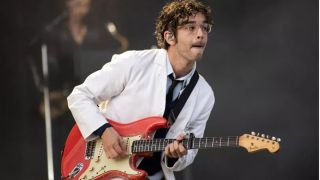 The 1975 frontman Matty Healy performing on stage in a white suit and black tie and playing a red electric guitar.