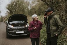 A mature couple discusses their situation in front of a broken down car on the road.