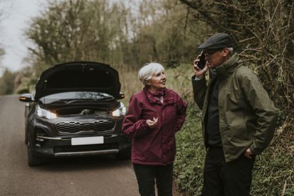 A mature couple discusses their situation in front of a broken down car on the road.