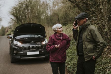 A mature couple discusses their situation in front of a broken down car on the road.