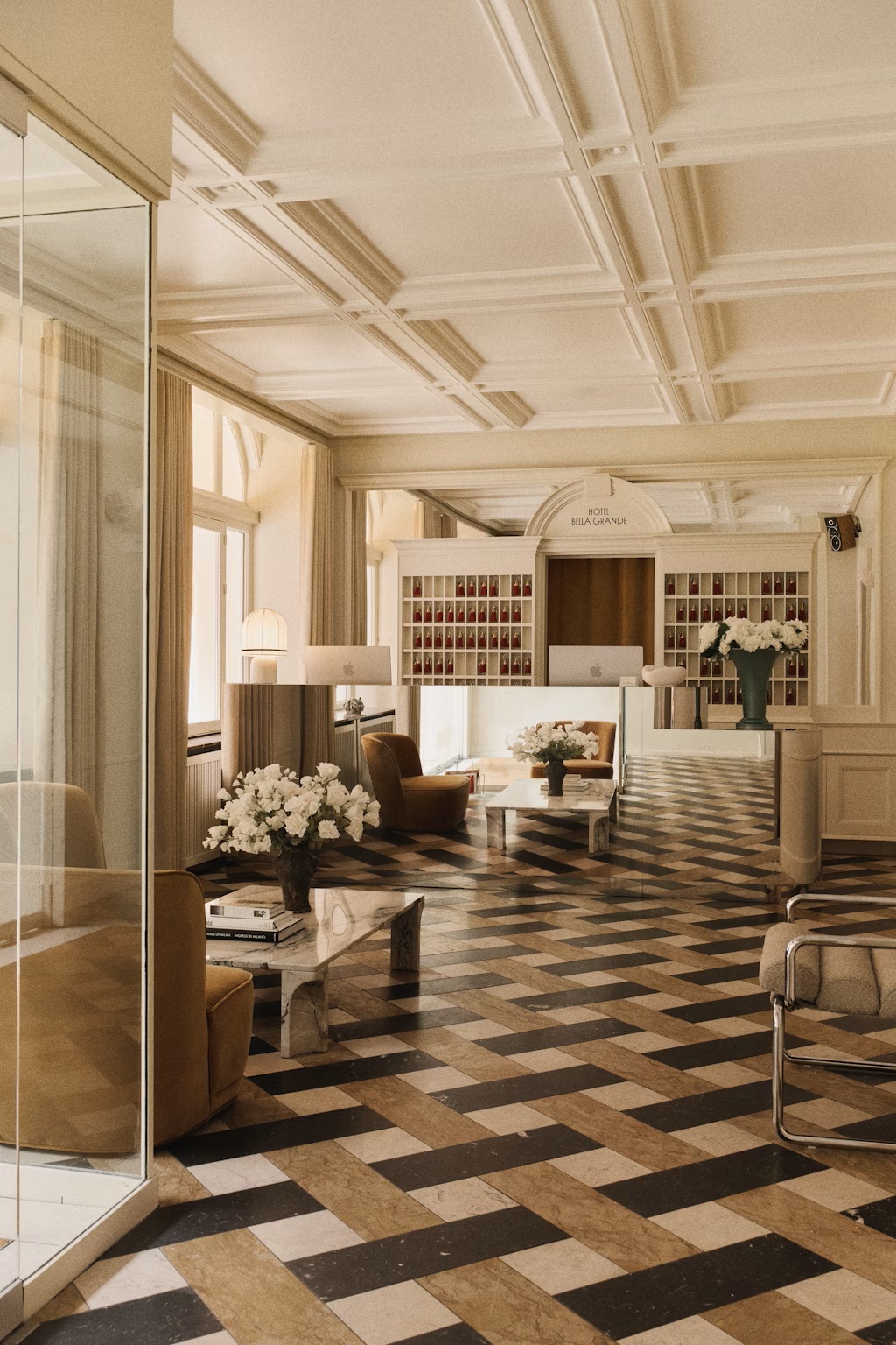A striking, retro-inspired hotel lobby features creme-tinted furnishings, including chrome and leather amrchairs, velvety sofas, mushroom-shaped Murano glass table lamps, and a theatrical reception, with checkered floors.