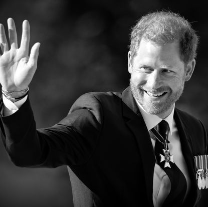 A black and white photo of Prince Harry wearing a suit and tie smiling and waving