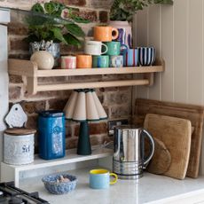 White kitchen cabinets with exposed brick walls, wooden shelf, stack of colourful mugs, green lamp and silver kettle.