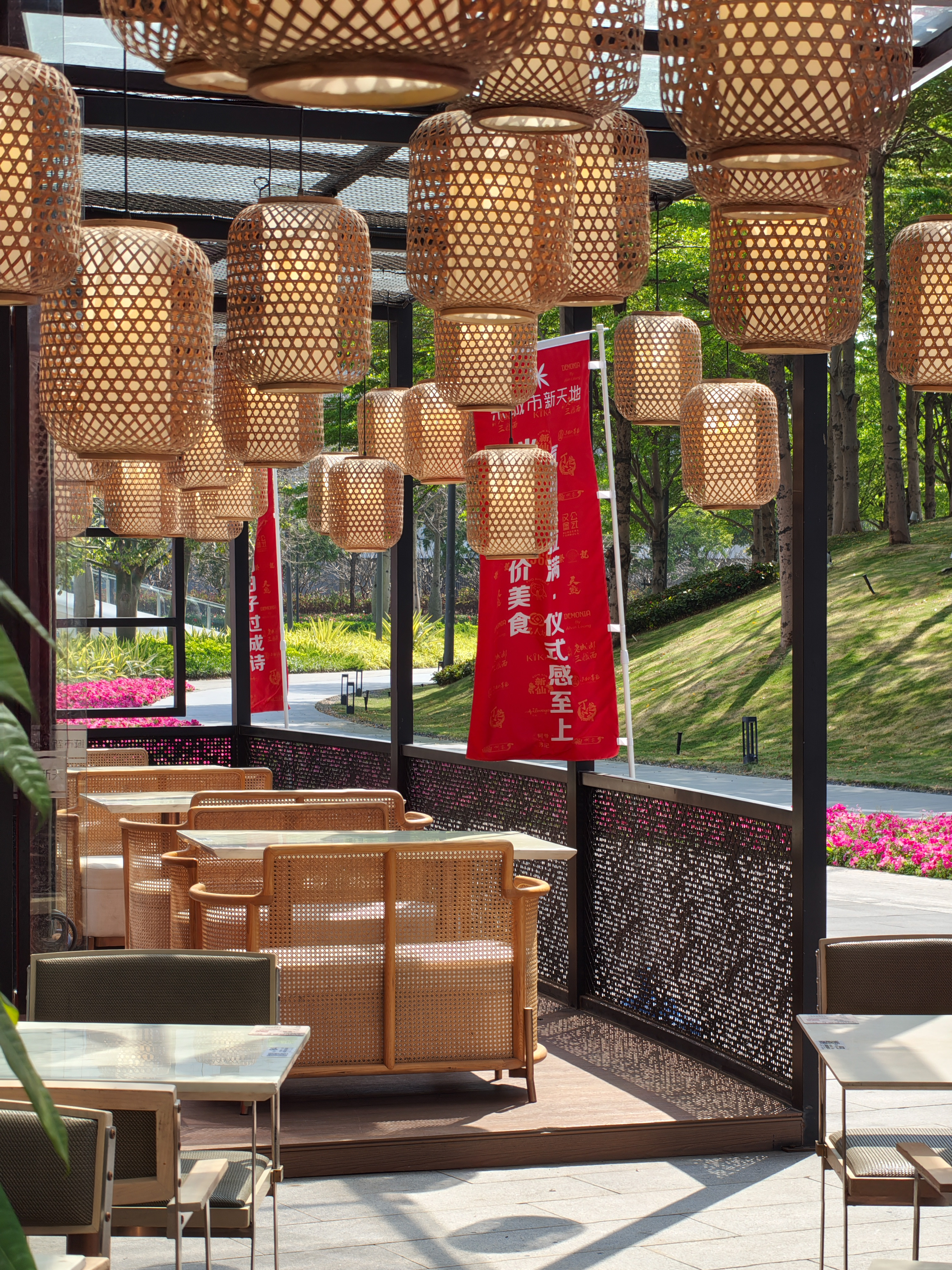 An outdoor patio area of a restaurant or cafe, viewed through several hanging woven bamboo lanterns that fill the upper half of the frame. Wicker sofas with light-colored cushions face white tables. In the background, vibrant red vertical banners with Chinese text stand near a lush green hillside and pink flower beds under bright sunlight.