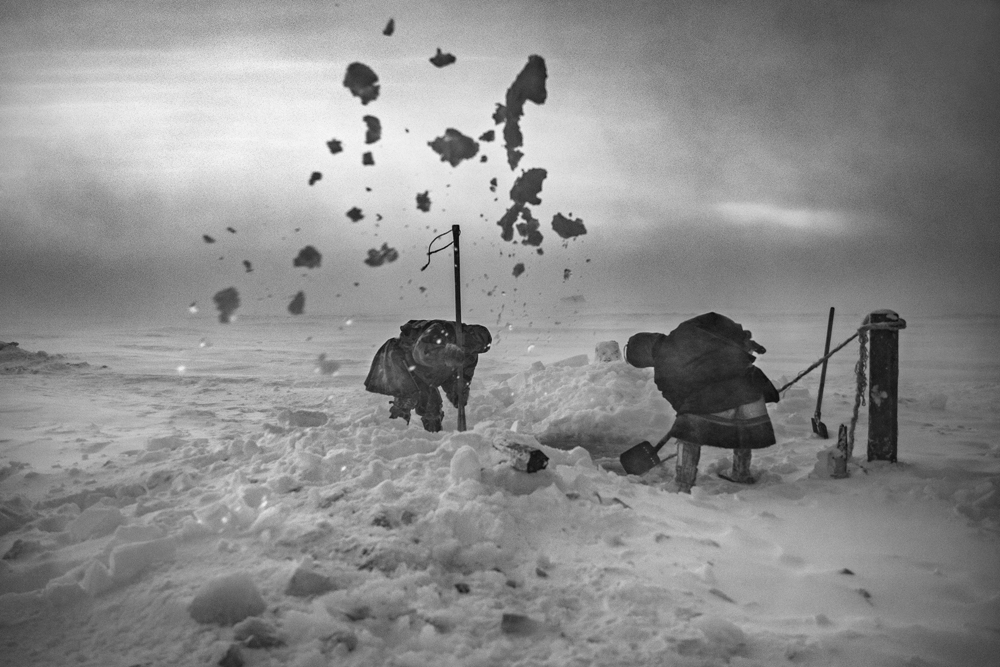 Two figures shovel snow under a cloudy sky, creating snowflakes that scatter in the wind, surrounded by a snowy landscape