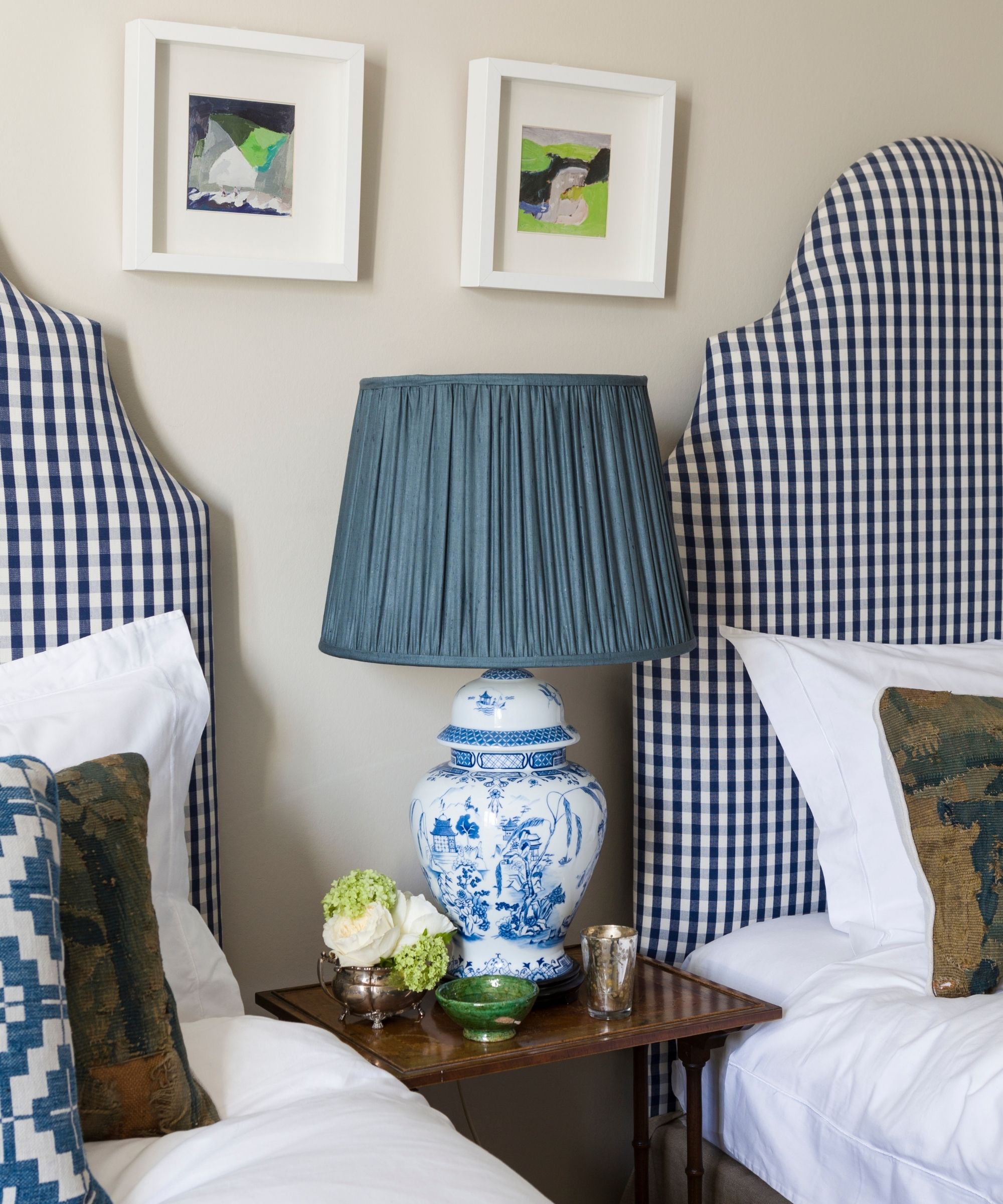 A twin bedroom with blue and white gingham headboards, white bedding, matching cushions, and a ceramic lamp with blue lampshade on a dark wood nightstand between the beds.