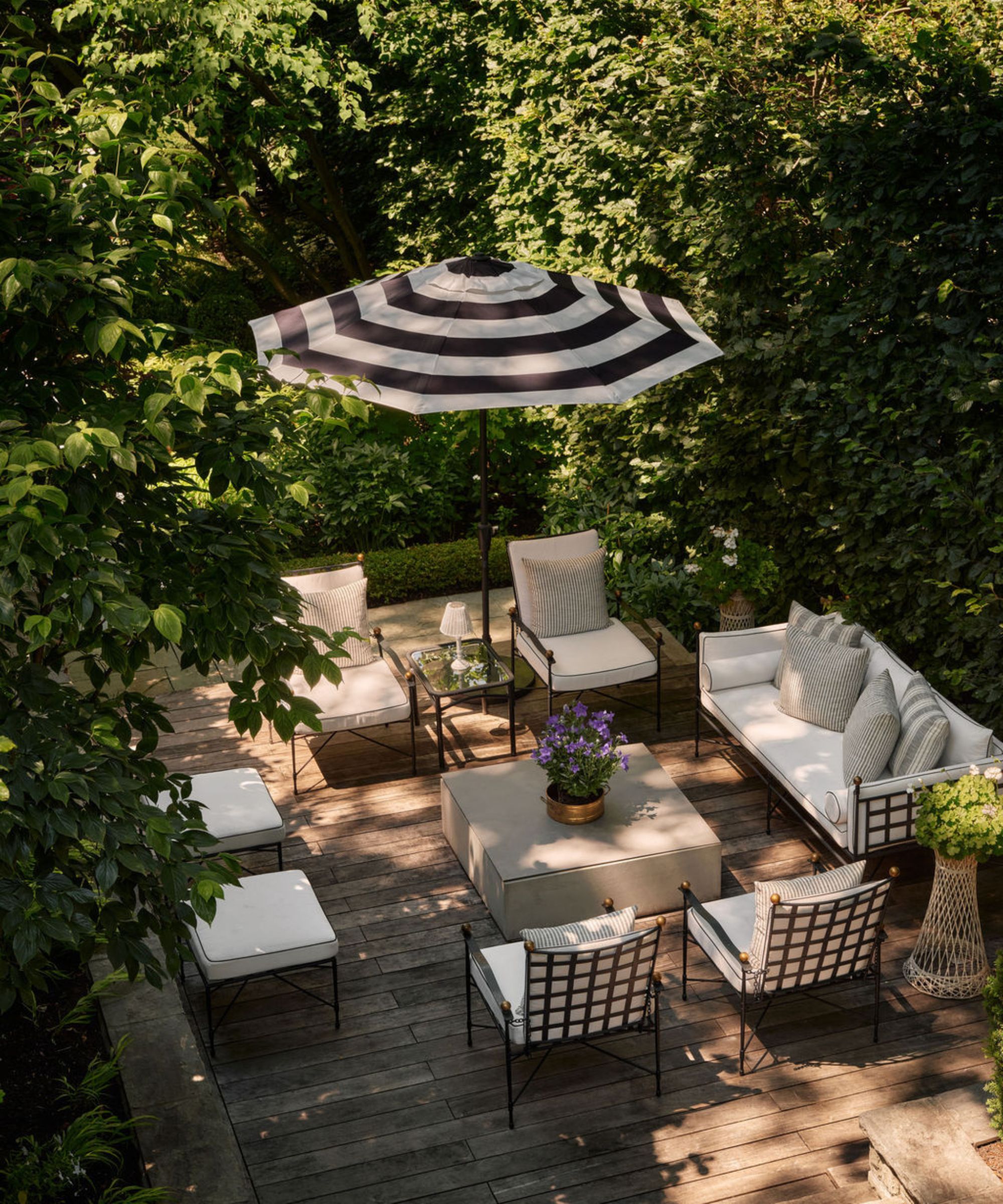 a small backyard terrace surrounded by trees with black and white antique furniture and a striped parasol