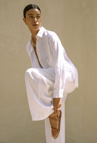 a photo of a model wearing a white linen shirt and trousers with tan flat sandals from the minimal sandal brand amanu