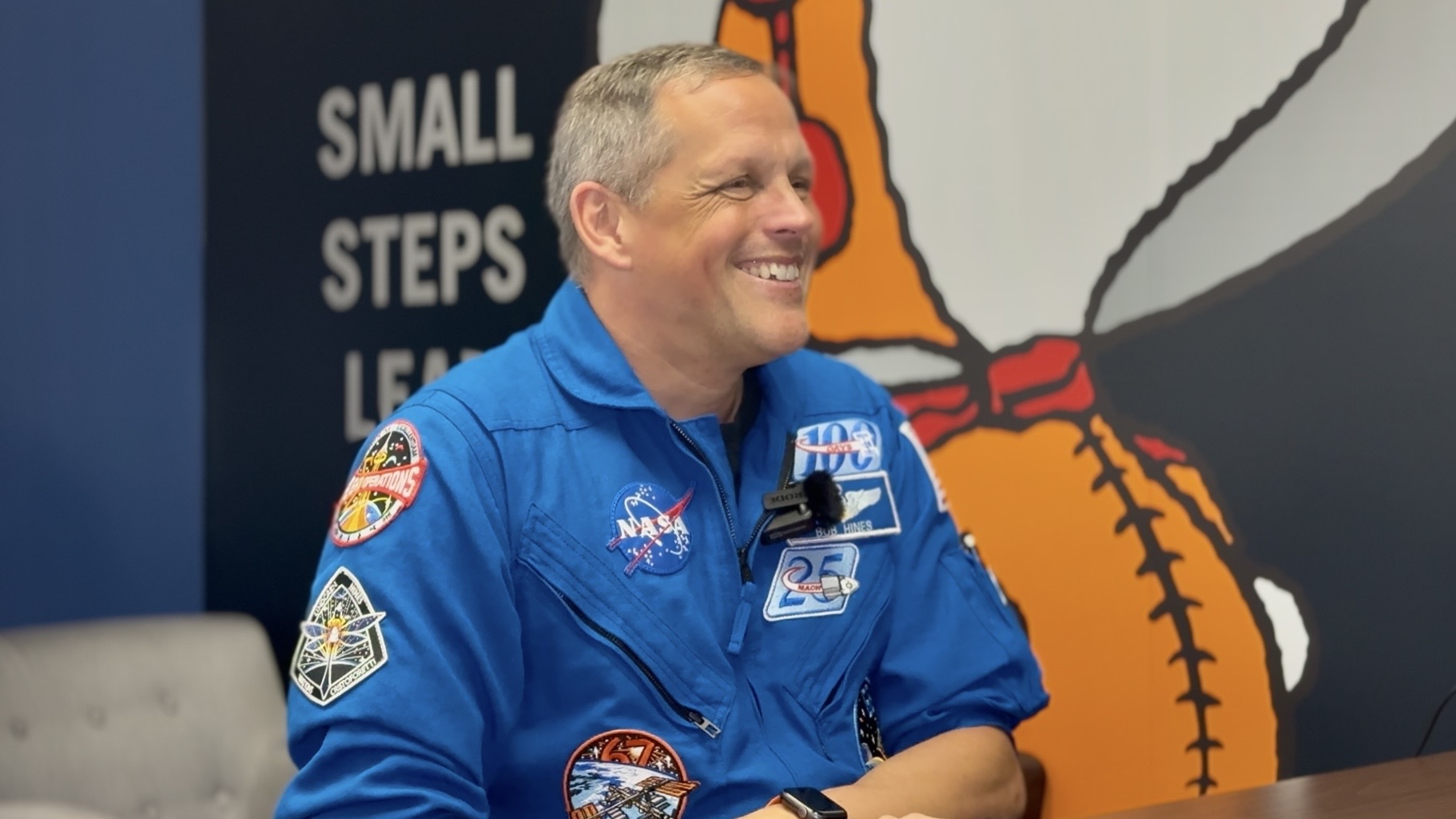 A man in a blue jumpsuit sits smiling in front of a wall with a giant snoopy astronaut painted on.