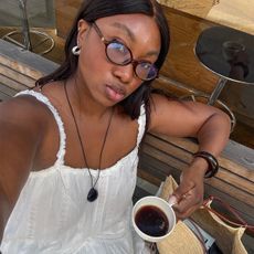 Influencer Nnenna wears a white lace dress and a long pendant necklace while holding a cup of coffee. 