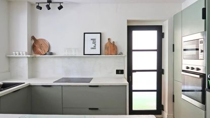 Learning how to microcement over tiles is so easy. Here is a kitchen with microcemented walls, a wall shelf with wall art and chopping boards on it, a white countertop with a gray cabinet, and a black PVC door
