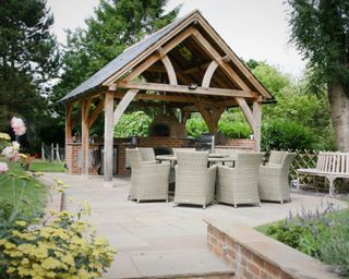 A timber-framed gazebo with a slate tiled roof, housing a brick outdoor kitchen with large round table and wicker chairs on a traditional paved patio.