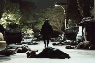 A dark, hooded figure walks through along a road littered with corpses