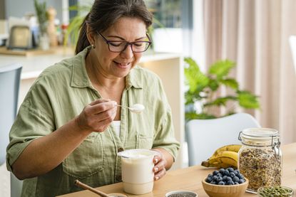 Woman holding spoon of Greek yoghurt, with a bowl of blueberries, a bowl of pumpkins, a container of oats and a bunch of bananas on the counter she's sitting at