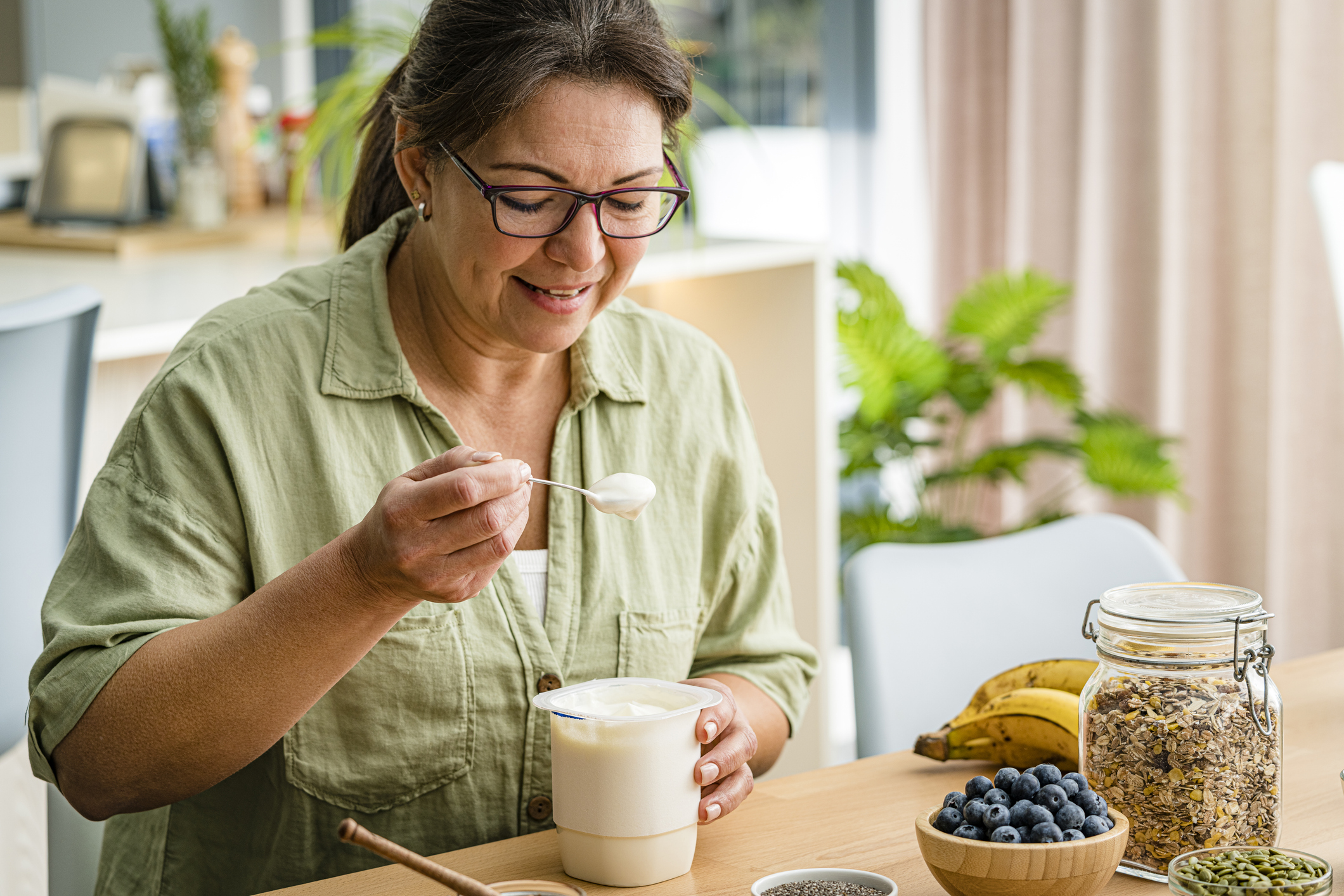 Woman holding spoon of Greek yoghurt, with a bowl of blueberries, a bowl of pumpkins, a container of oats and a bunch of bananas on the counter she's sitting at